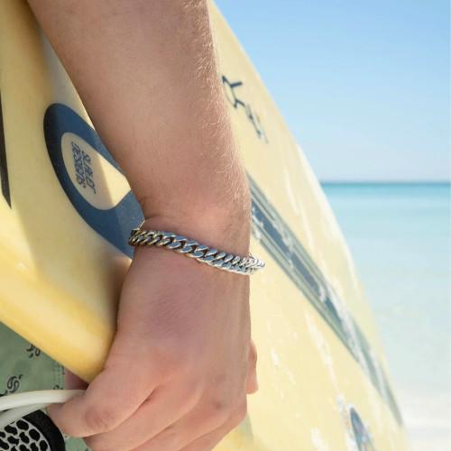 Man on the beach holding a surfboard, wearing the 100% waterproof Cuban mesh stainless steel bracelet.